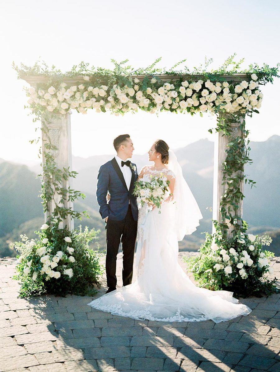 haute couture bridal gown with long sleeves and navy groom suit with elegant outdoor wedding arch backdrop